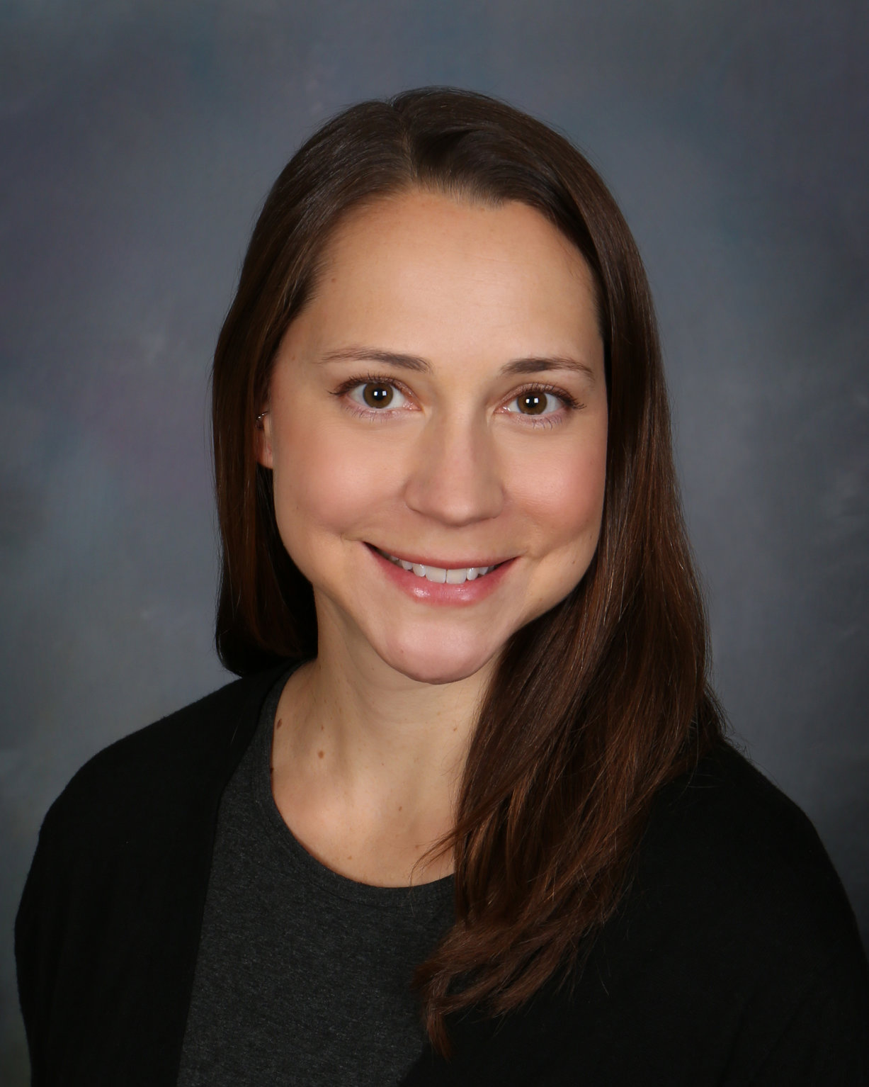 A woman with long brown hair smiles at the camera. She is wearing a dark top and a black cardigan, with a neutral gray background behind her.