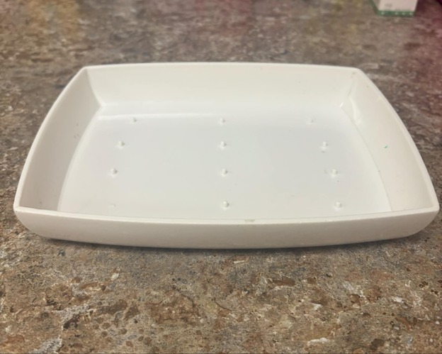 A white rectangular soap dish with small raised bumps on the bottom is placed on a brown speckled countertop.