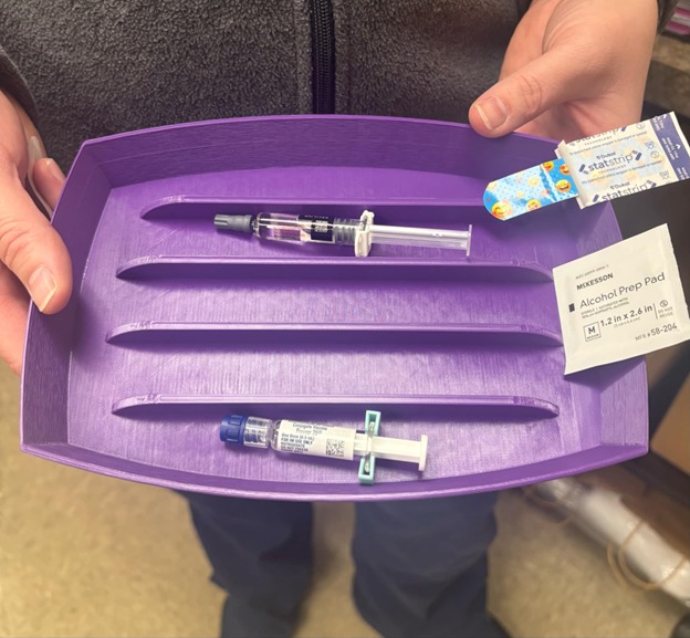 A person holds a purple tray containing two syringes, an alcohol prep pad, and a decorated adhesive bandage in a medical setting.