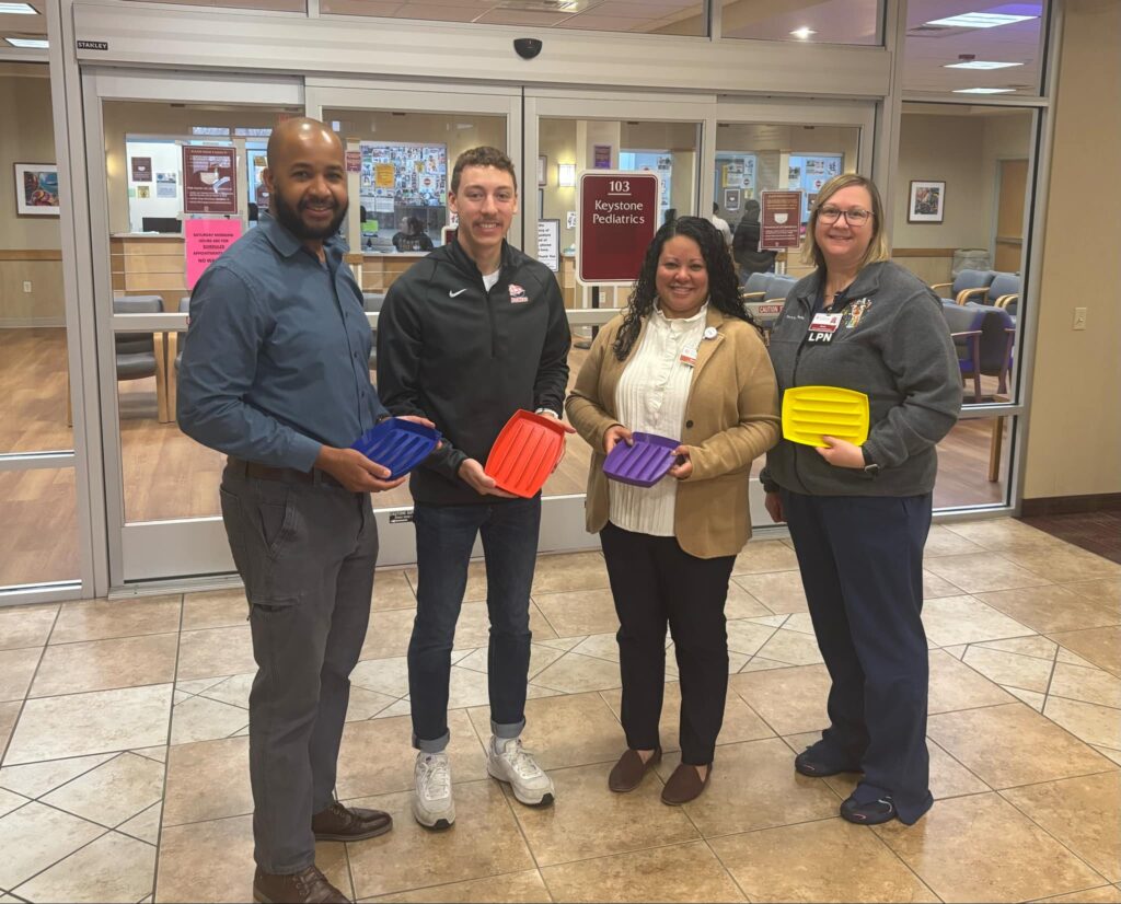 Four people standing indoors in front of glass doors, each holding a colorful plastic tray (blue, red, purple, yellow), and smiling at the camera. The setting appears to be a clinic or office environment.