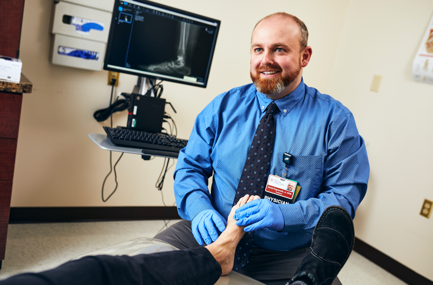 A smiling male doctor in blue gloves examines a patient's foot in a medical office, with an X-ray image of the foot displayed on a computer monitor in the background.
